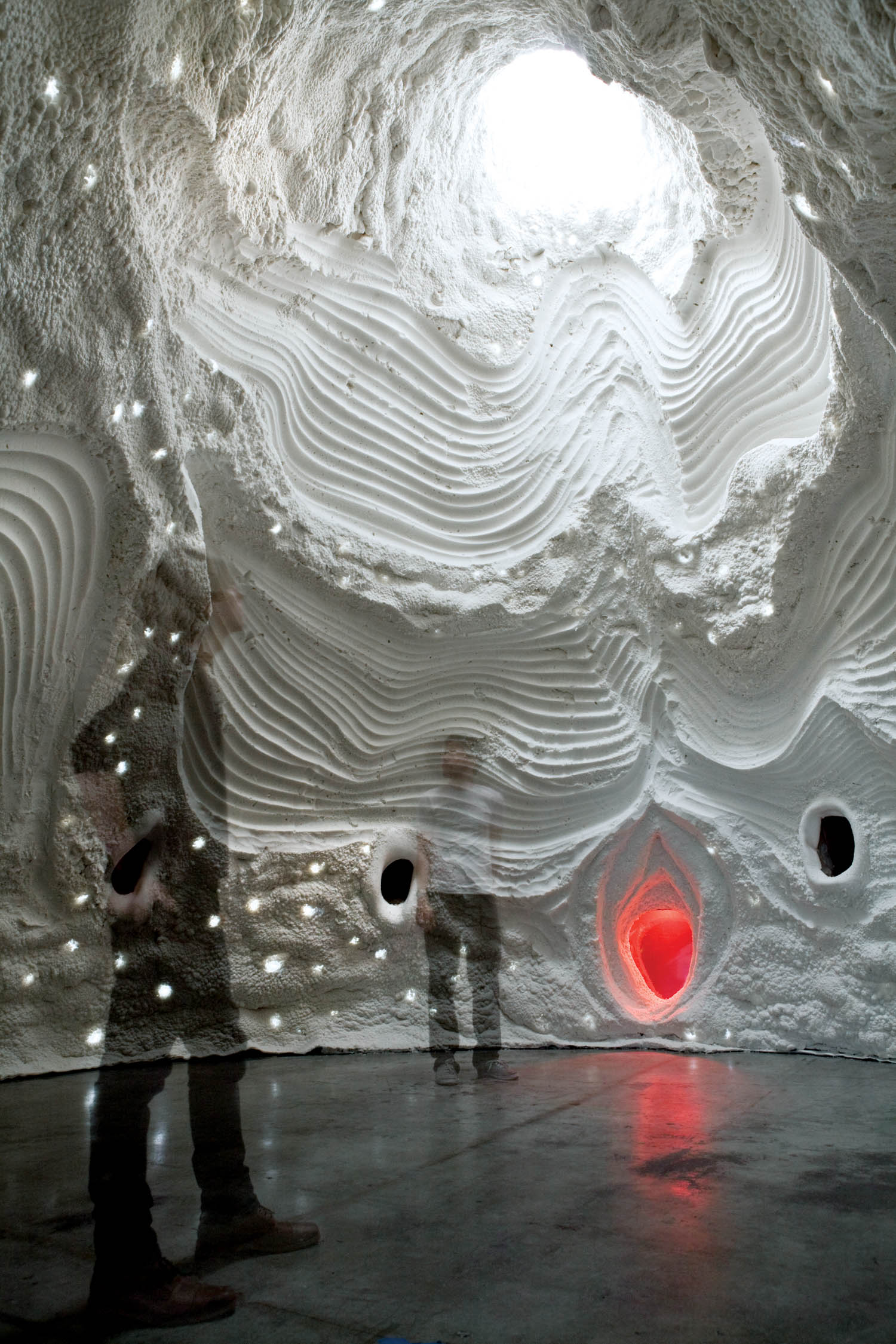 Two people stand inside a cave-like room with textured white walls, a skylight above, dotted light spots, and a glowing red feature set into one wall.