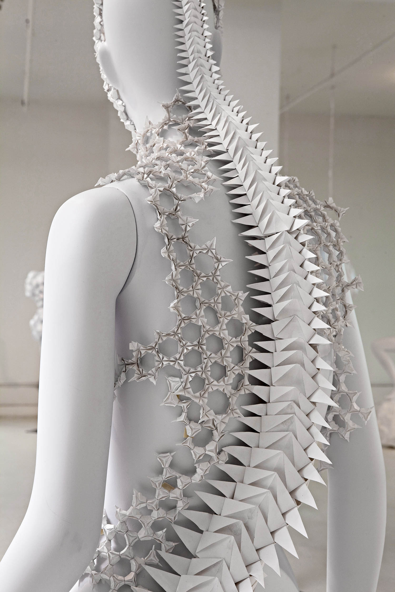 White mannequin wearing an intricate, geometric, spiked back piece made of interlocking paper or plastic shapes in a minimalist gallery setting.