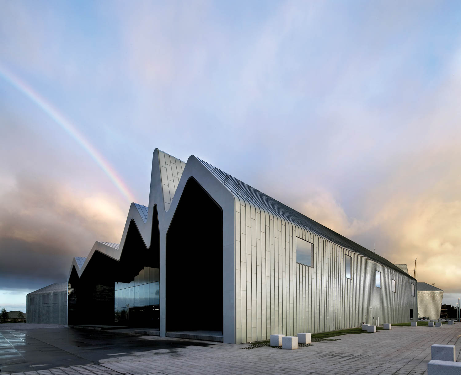 Modern industrial building with a zigzag roof, clad in metallic panels, under a cloudy sky with a visible rainbow in the background.