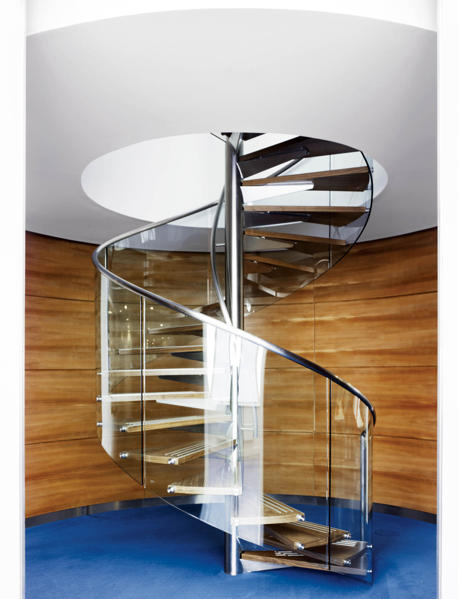 Modern spiral staircase with glass sides and metal railings, set against curved wooden walls and a blue floor, leading up through a circular ceiling opening.