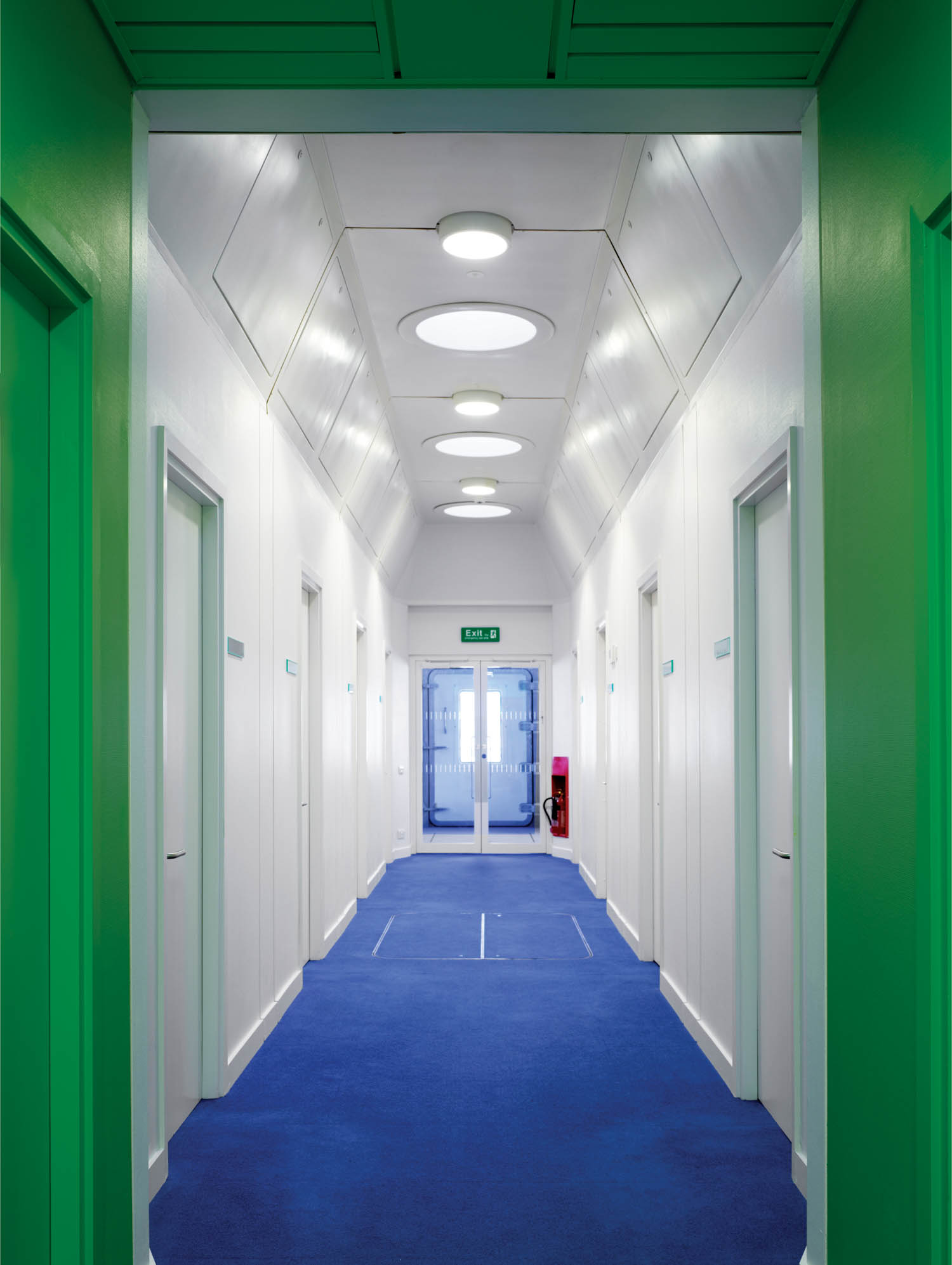 A brightly lit hallway with blue carpet, white walls, green door frames, and several closed doors leading to a glass exit at the end.