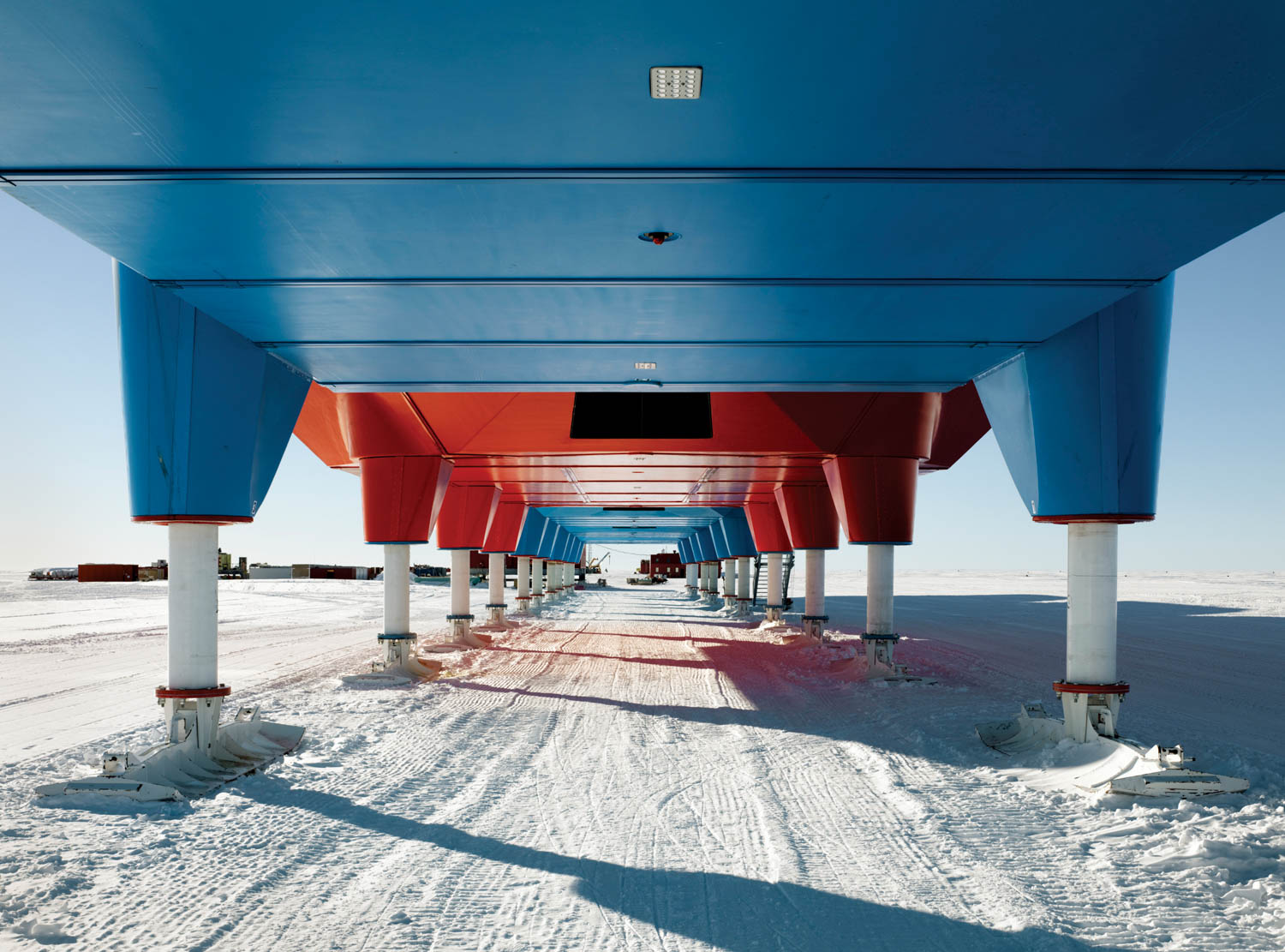 View under an elevated blue and red building on snowy ground, with clear sky and distant structures visible in the background.