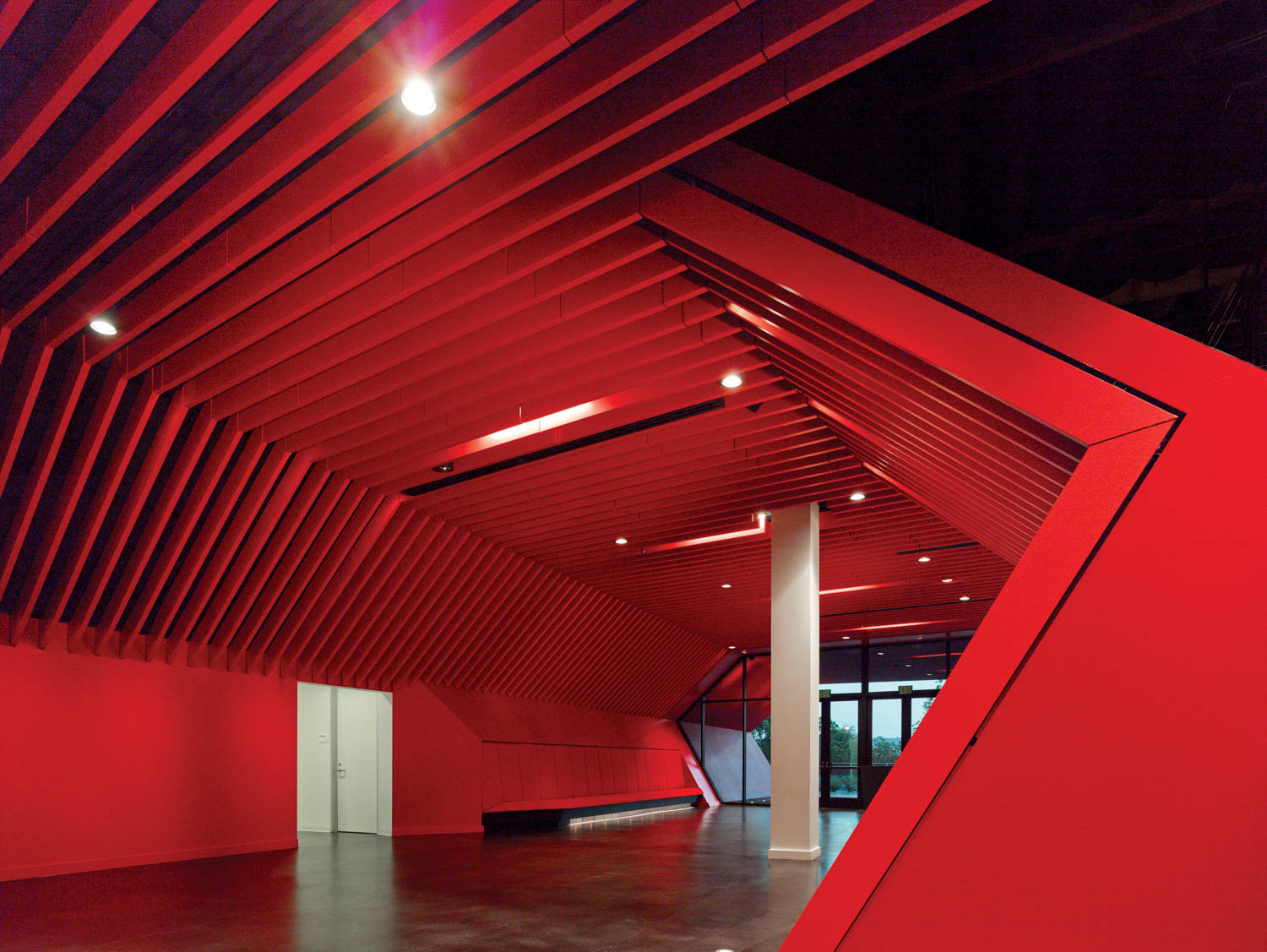 A modern interior space features angular red walls and ceiling panels, a white pillar, dark flooring, and a glass entrance in the background.