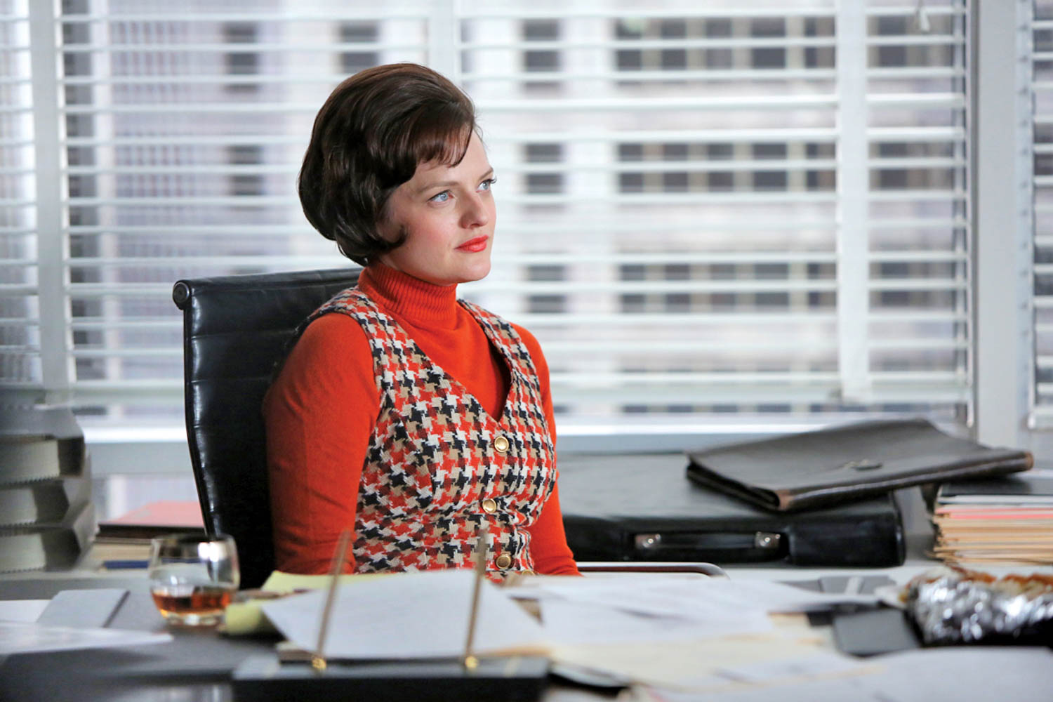 Woman sits at a cluttered office desk, wearing a red turtleneck and patterned dress. She faces slightly to the side with a neutral expression. Blinds and city buildings are visible behind her.