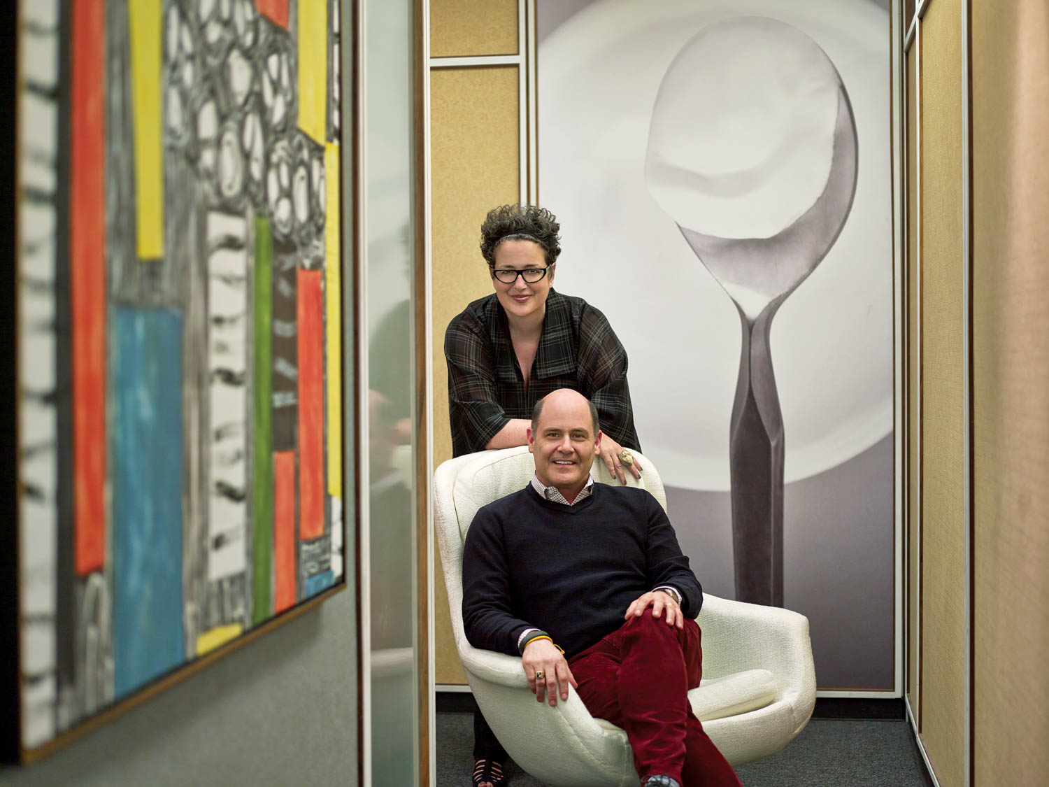 A woman stands behind a seated man in a modern office hallway with colorful abstract art and a large spoon artwork on the wall.