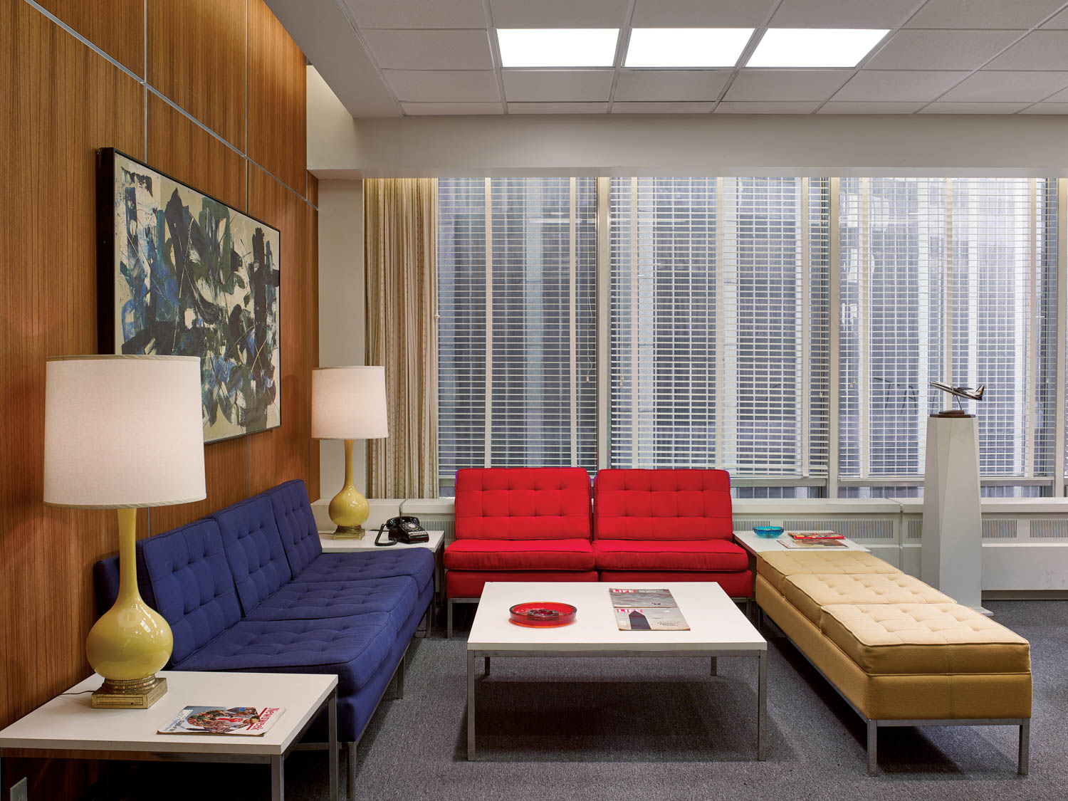 A mid-century modern office lounge with blue, red, and yellow sofas, two yellow lamps, a coffee table, magazines, and large windows with blinds.