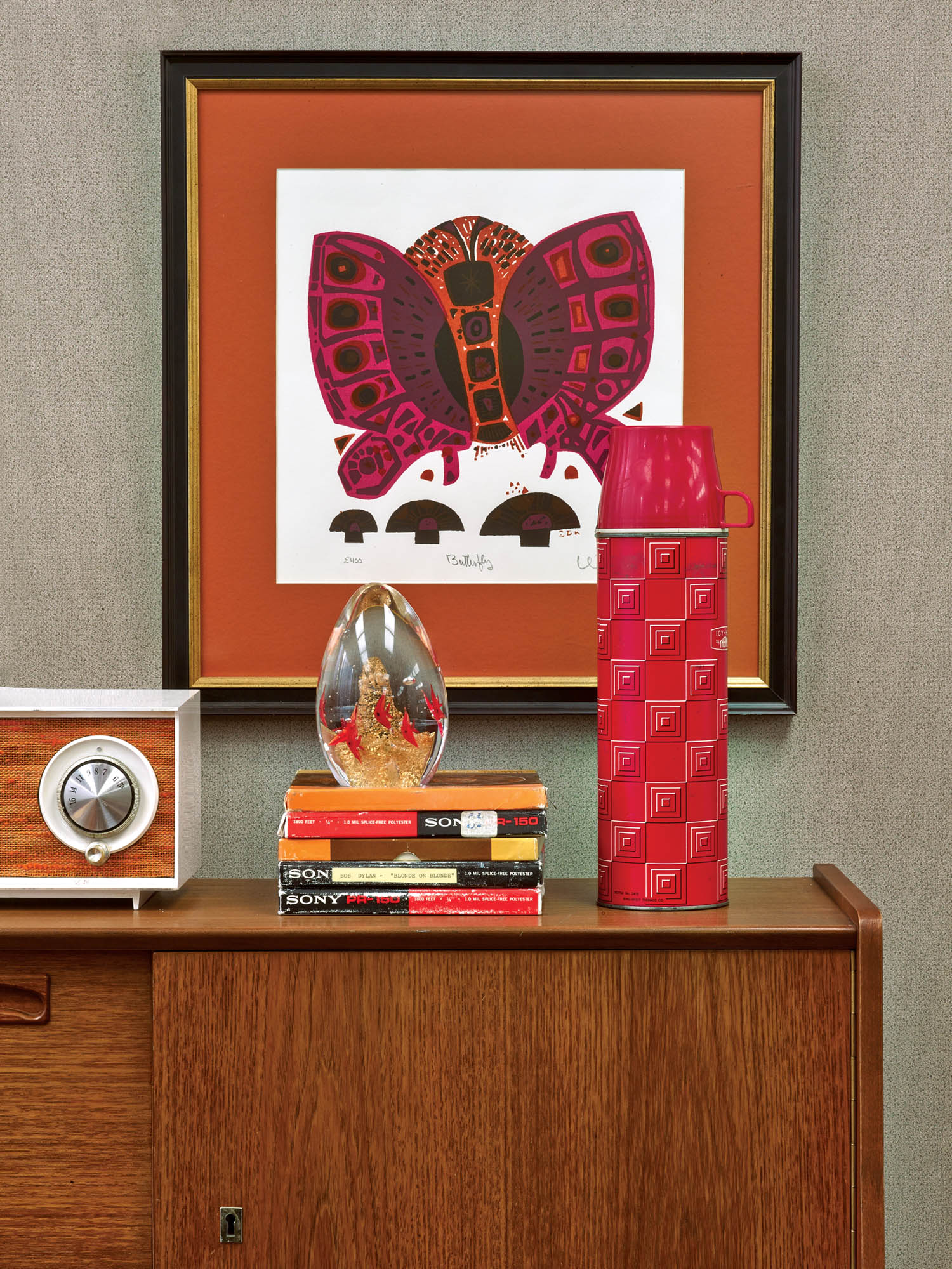 A framed butterfly print hangs above a wooden cabinet with a radio, glass ornament, stacked books, and a tall red thermos on top.