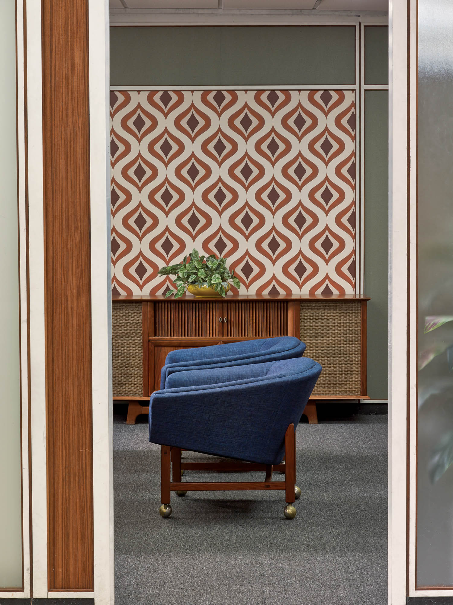 A blue chair faces a wooden cabinet with a potted plant, set against a wall with geometric brown and white wallpaper in an office space.