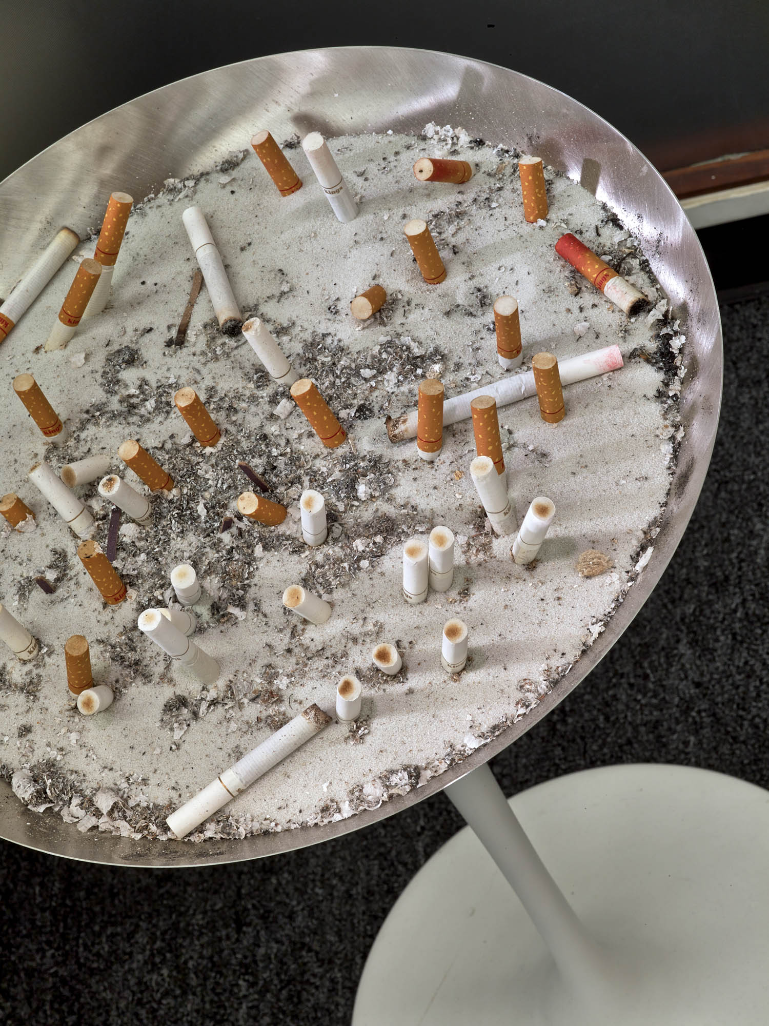A round metal ashtray filled with cigarette butts and ashes, standing on a pedestal against a dark background.