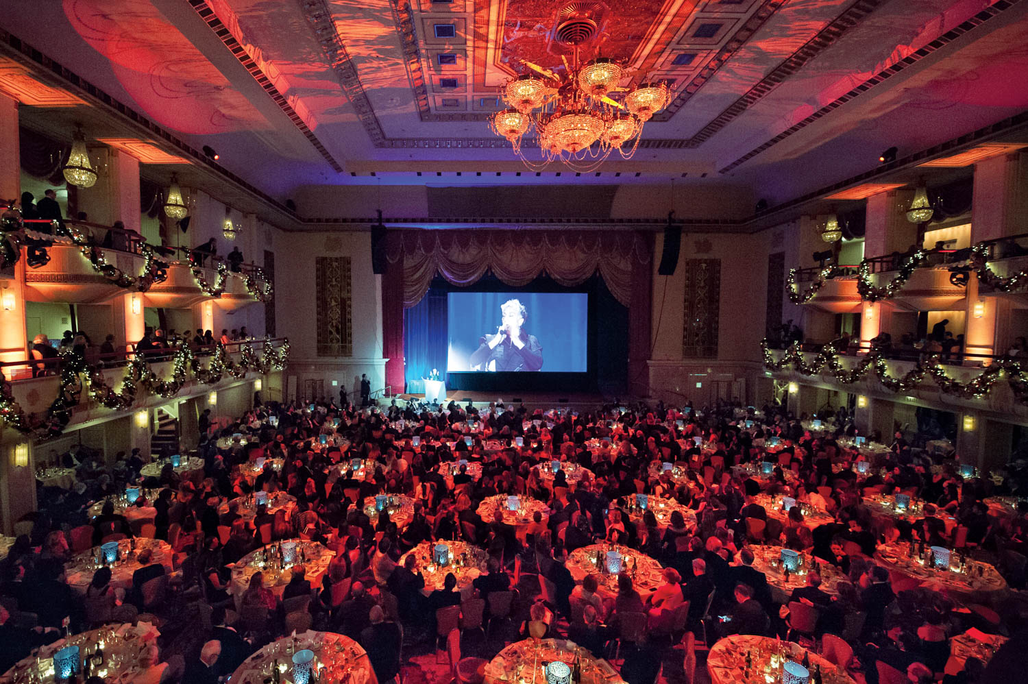 A large, ornate ballroom filled with round dining tables and guests facing a stage with a screen displaying a person speaking.