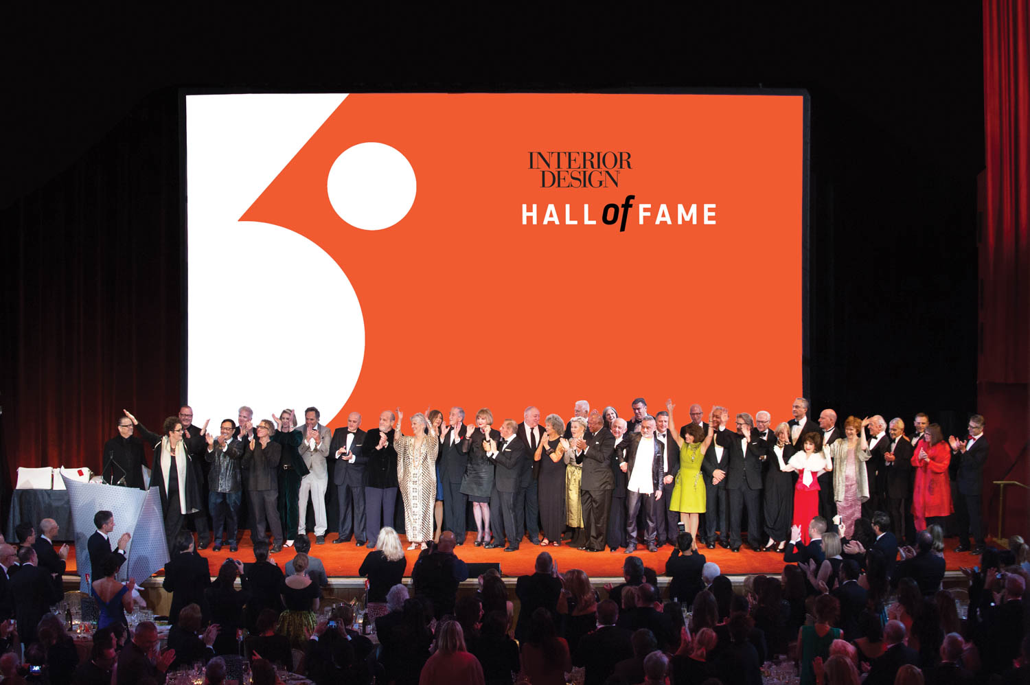 A large group of people stands on stage in front of a screen that reads “Interior Design Hall of Fame,” while an audience sits and watches.