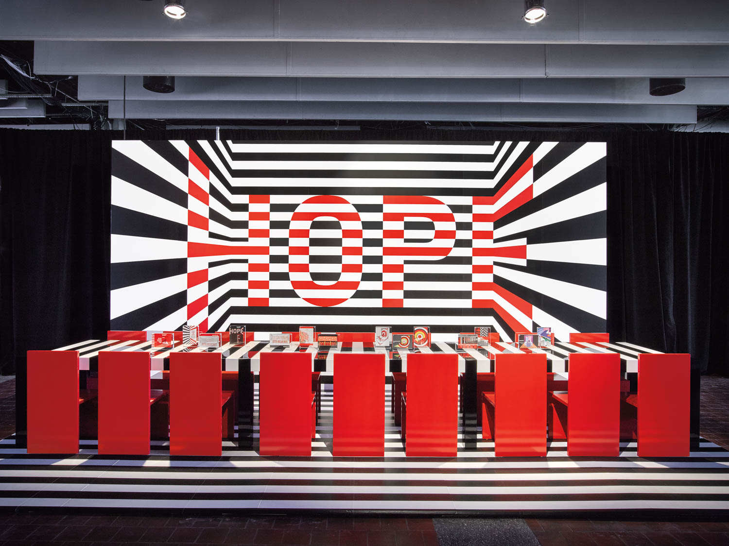 A modern room with red tables and chairs faces a black-and-white striped wall featuring the large word “HOPE” in bold red letters.