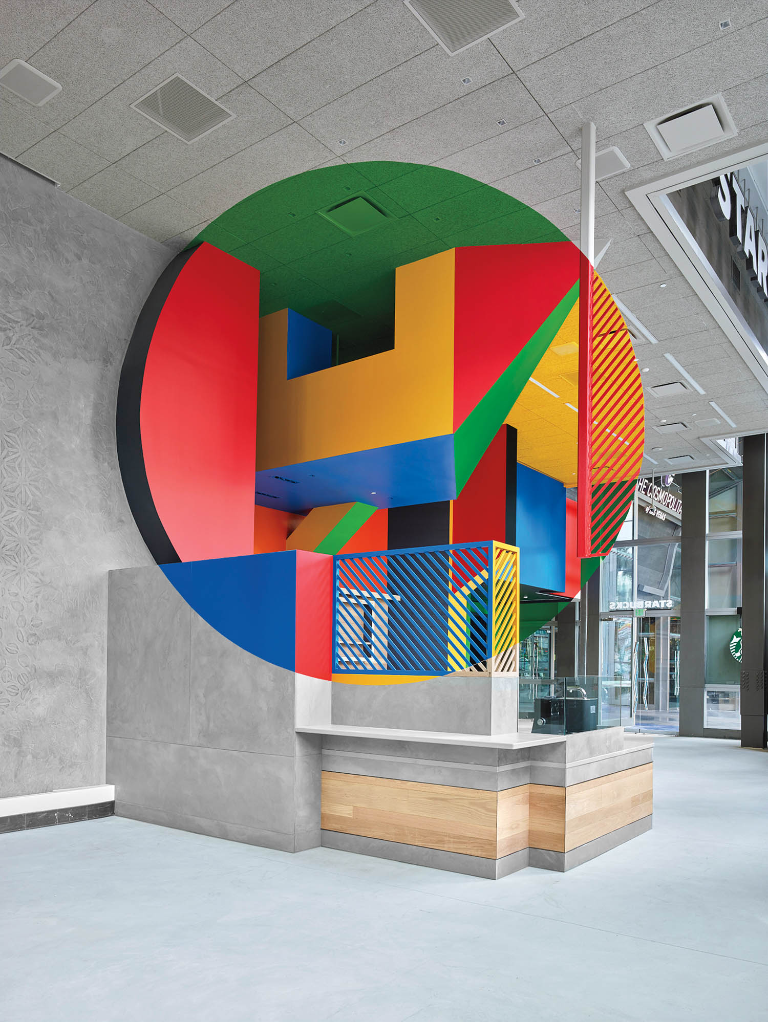 A colorful geometric mural with bold shapes and lines decorates a modern indoor space with a wood and glass counter in the foreground.