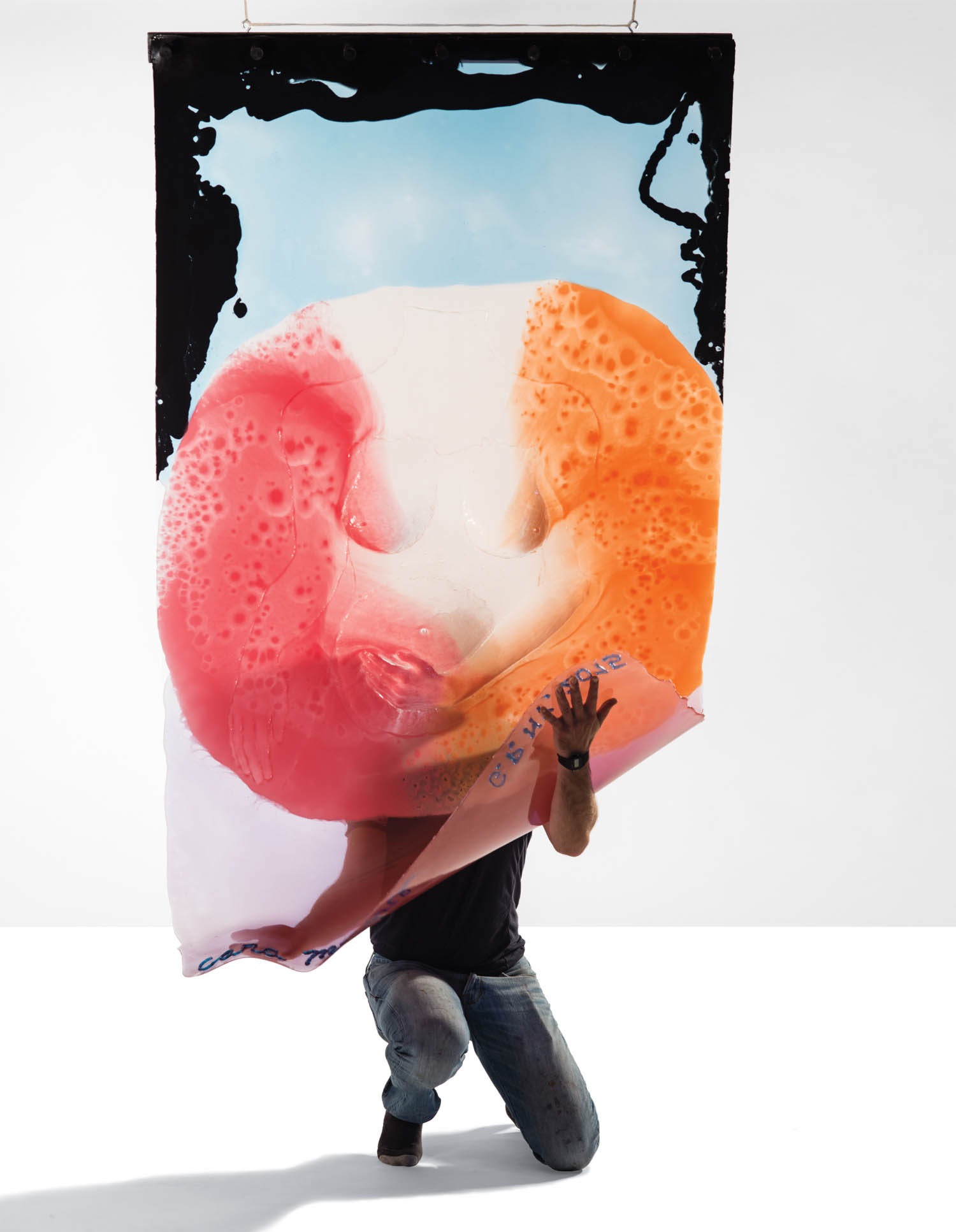 A person kneels, holding up a large, colorful, abstract artwork featuring red, orange, and white foamy shapes against a plain background.