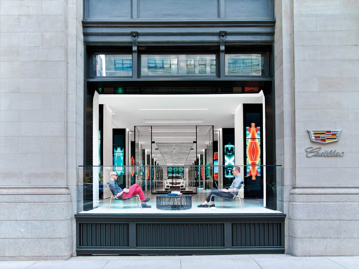 Two people sit in chairs facing each other in a modern Cadillac showroom with large glass windows and colorful neon lights inside.