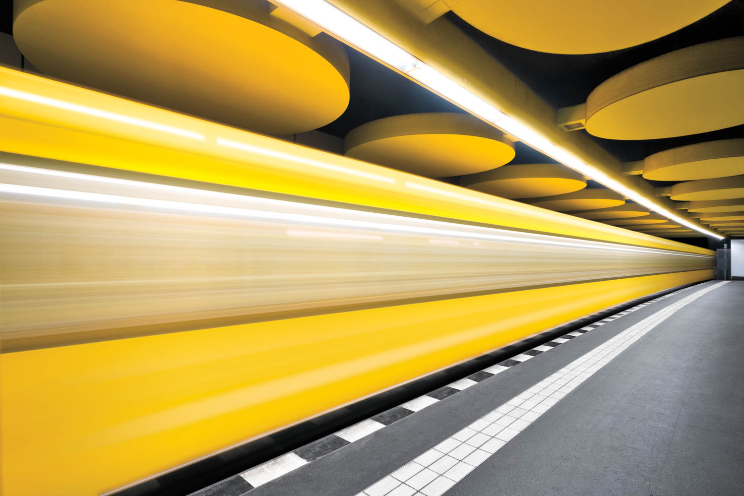 A yellow train moves quickly through a modern subway station, with circular ceiling features and a tiled platform in view.