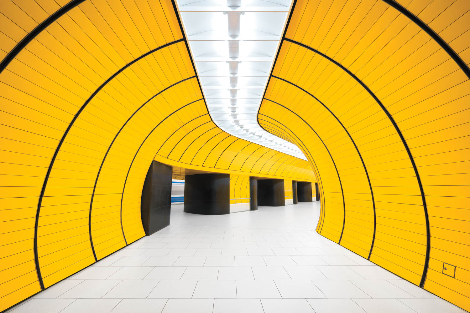 A modern underground station with curved yellow walls, black accents, and a white tiled floor under a bright ceiling.