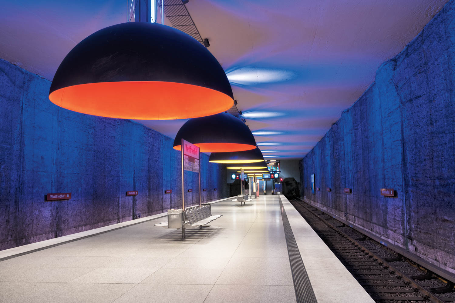 Modern subway platform with large black and orange dome lights, blue-lit textured walls, empty benches, and train tracks on the right side.