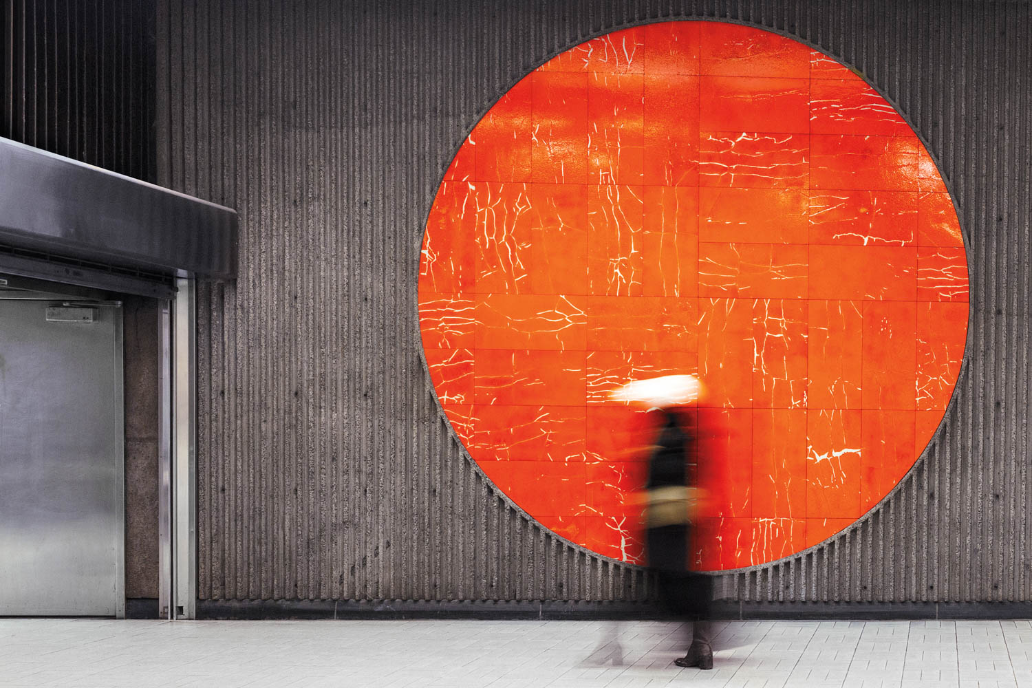 A large textured orange circle on a gray wall behind a blurred figure walking in an indoor public space.