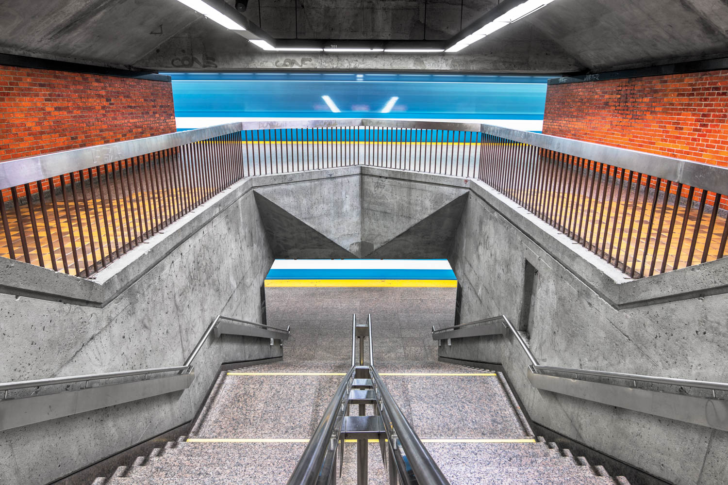 Concrete stairs and railings lead down to a subway platform with a moving blue train and brick walls in the background.