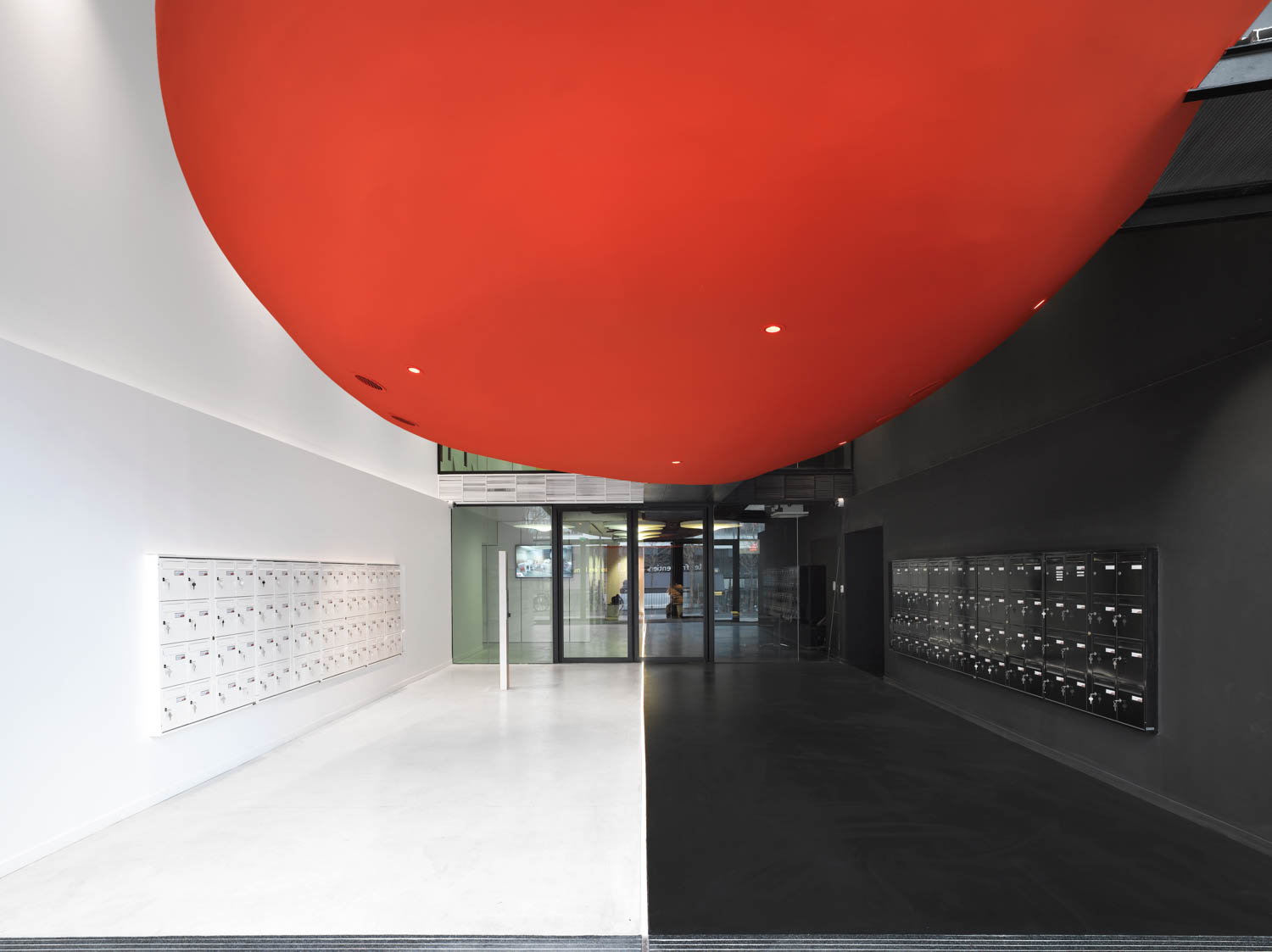 Modern building lobby with a large red ceiling feature, white and black floors, and rows of mailboxes mounted on both walls. Glass entry doors are visible at the far end.