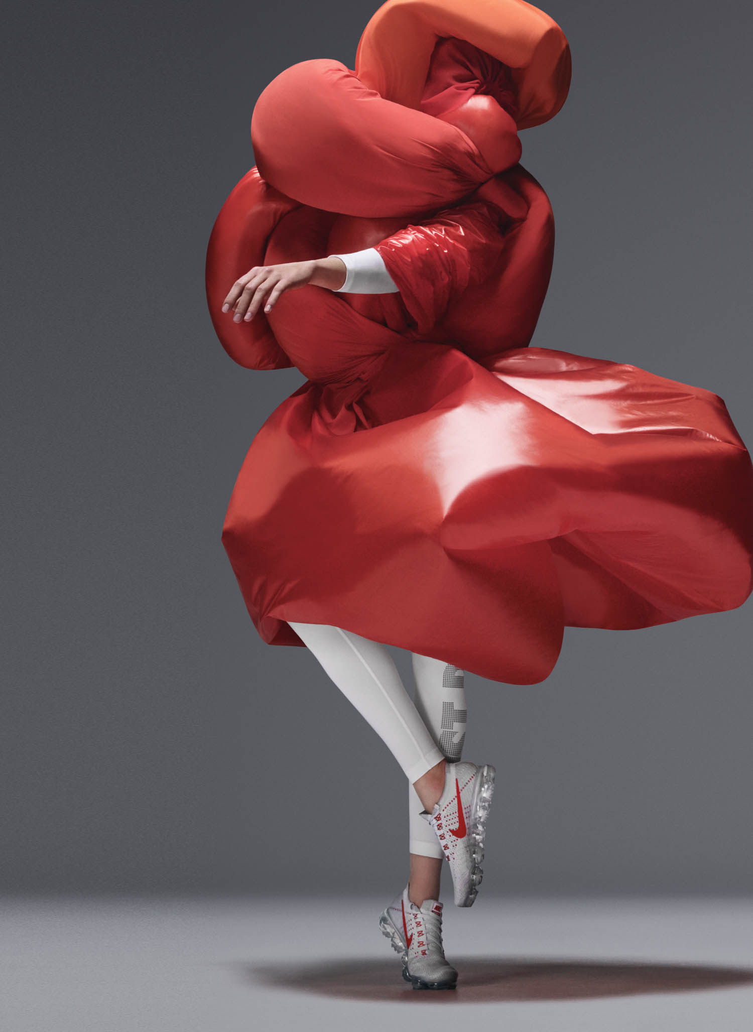 A person wearing a billowing red outfit and white leggings stands on one foot with arms crossed, wearing white sneakers with red accents against a gray background.