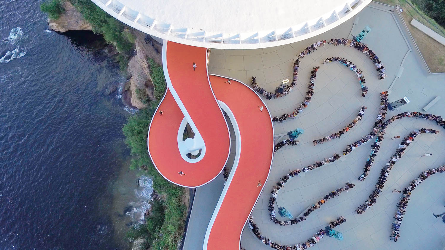 Aerial view of a curved red walkway next to a crowd of people lined up in winding rows on a light-colored plaza by the water.