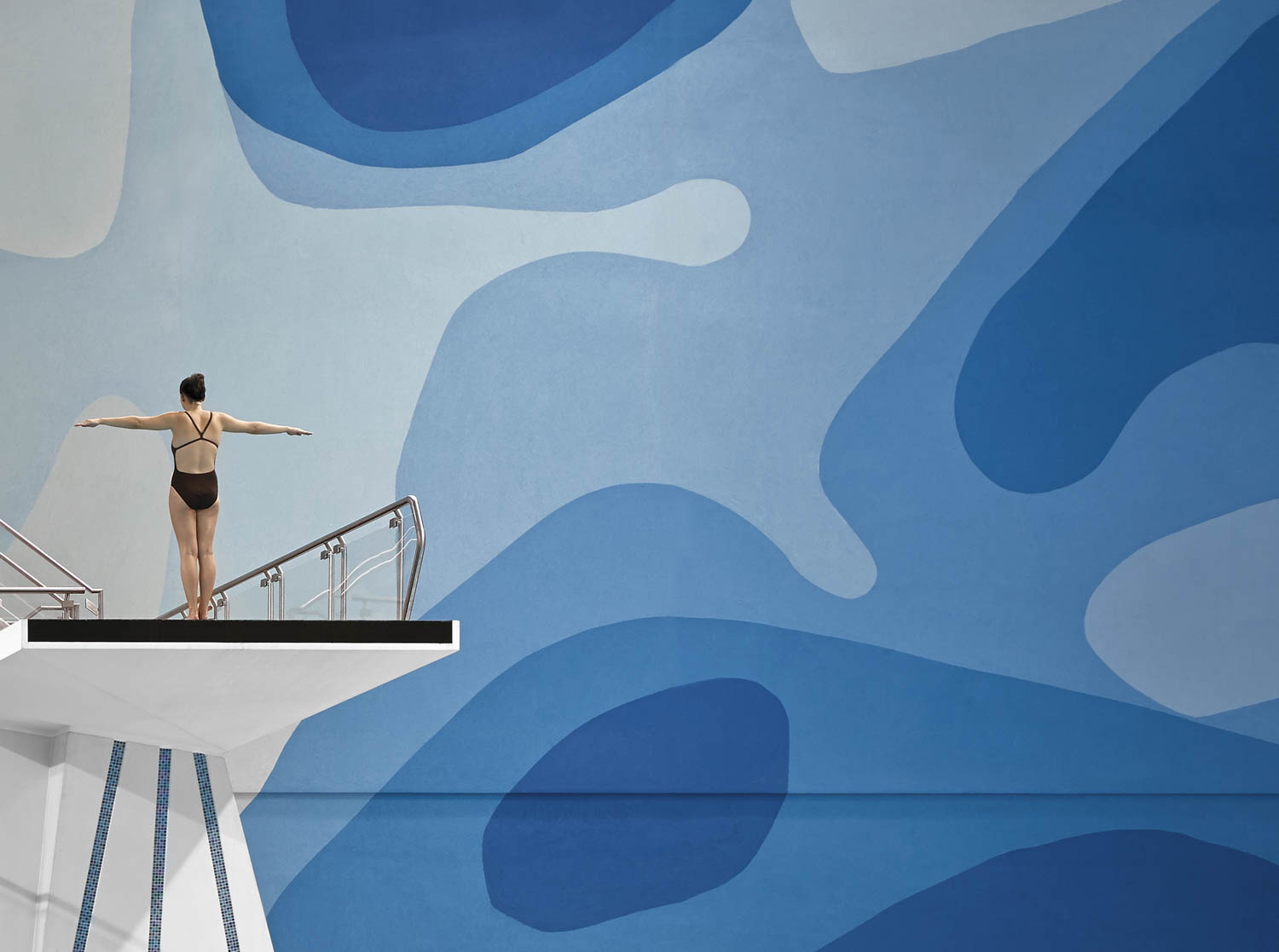 A person in a swimsuit stands with arms outstretched on a diving platform in front of a large wall with abstract blue patterns.