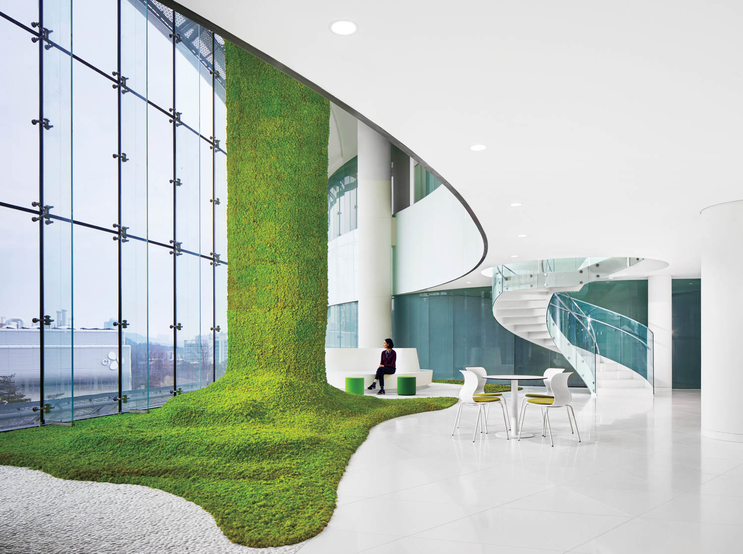 Modern office space with white floors, a curved staircase, floor-to-ceiling windows, and a large green moss installation extending from the wall onto the floor.