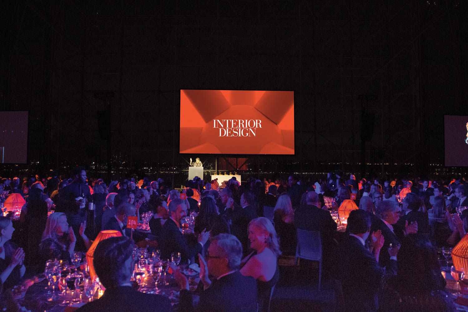 A large audience seated at round tables attends an event with a stage and a screen displaying “Interior Design” in white text on a red background.