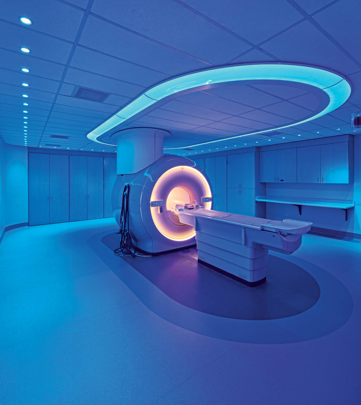 MRI machine in a modern, blue-lit hospital room with cabinets and a counter along the wall.