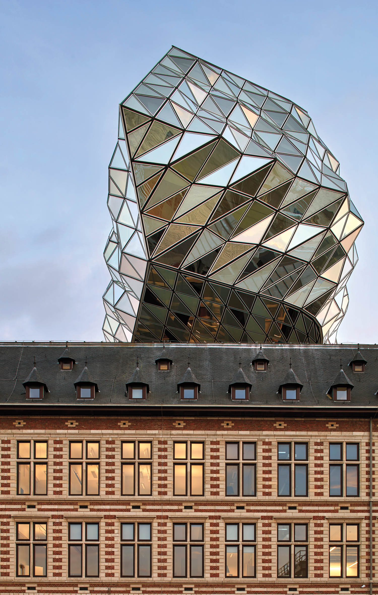 Modern glass geometric structure sits atop a traditional brick building with rectangular windows, set against a clear sky.