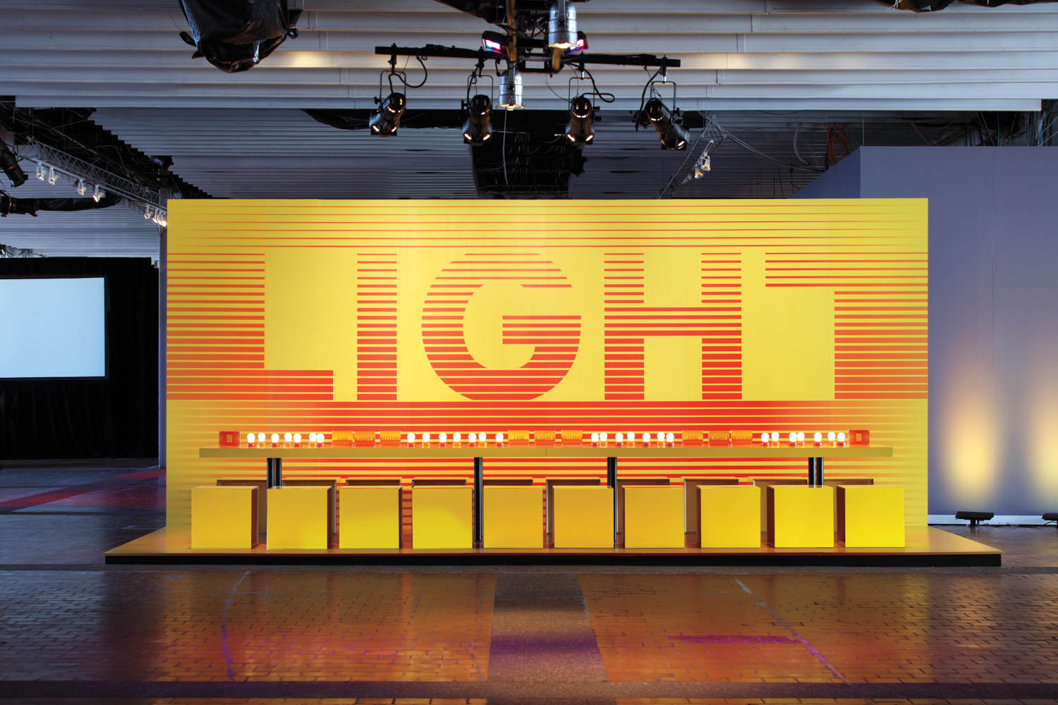 A display with a yellow wall featuring the word “LIGHT” in large letters, illuminated bulbs, and yellow blocks arranged in front.