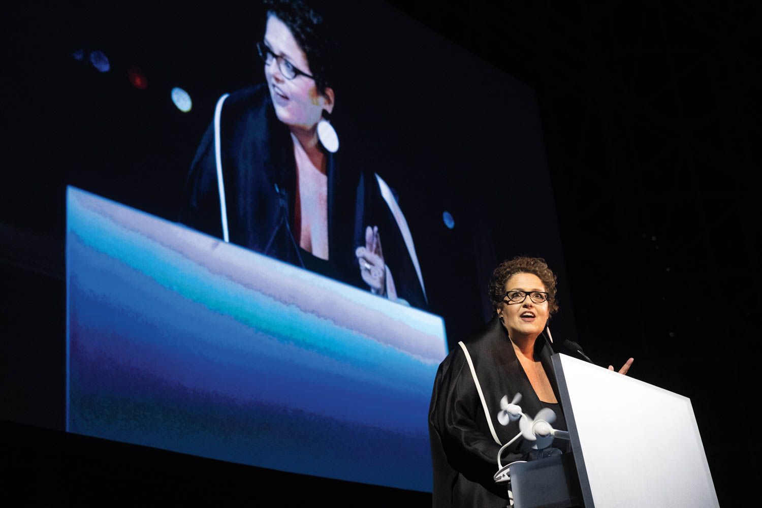 A woman wearing glasses speaks at a podium with a microphone, with her image projected on a large screen behind her.