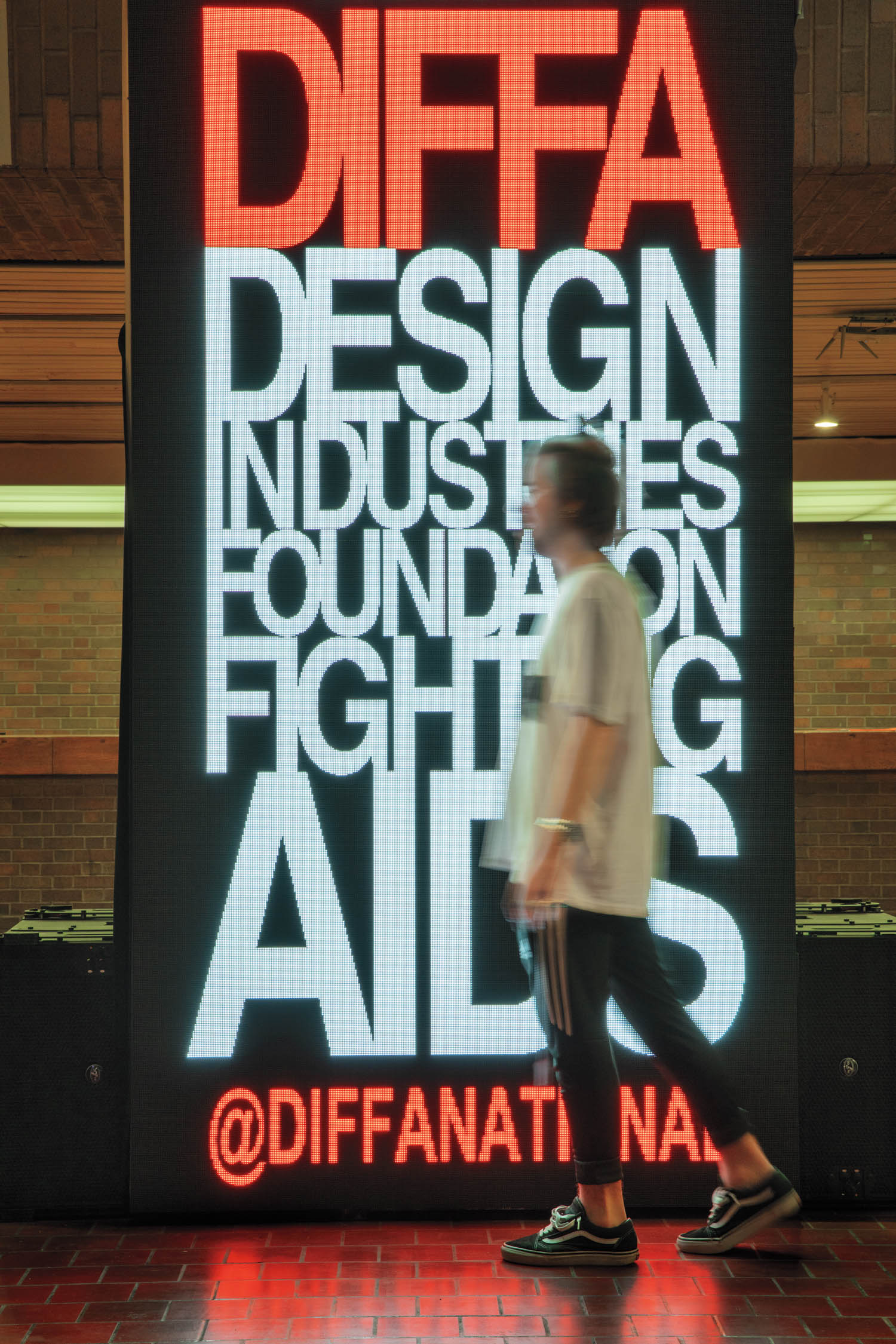 A person walks past a large illuminated sign that reads, “DIFFA: Design Industries Foundation Fighting AIDS @DIFFANATIONAL.”.