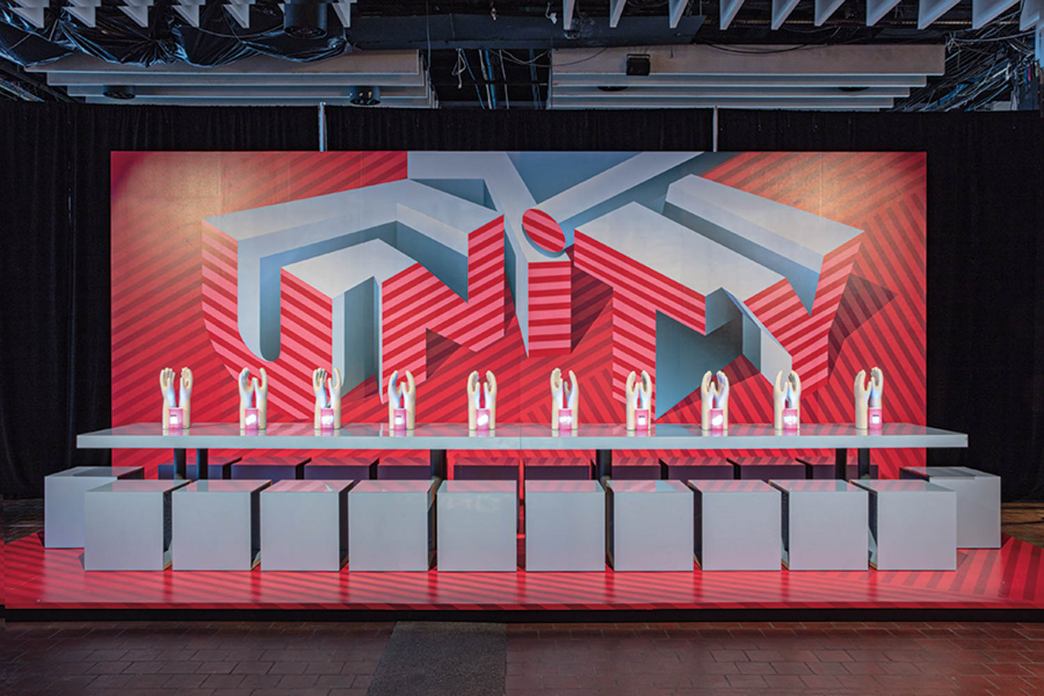 A row of twelve trophies is displayed on a long table in front of a geometric red and gray UNITY backdrop, with white cubes beneath the table.