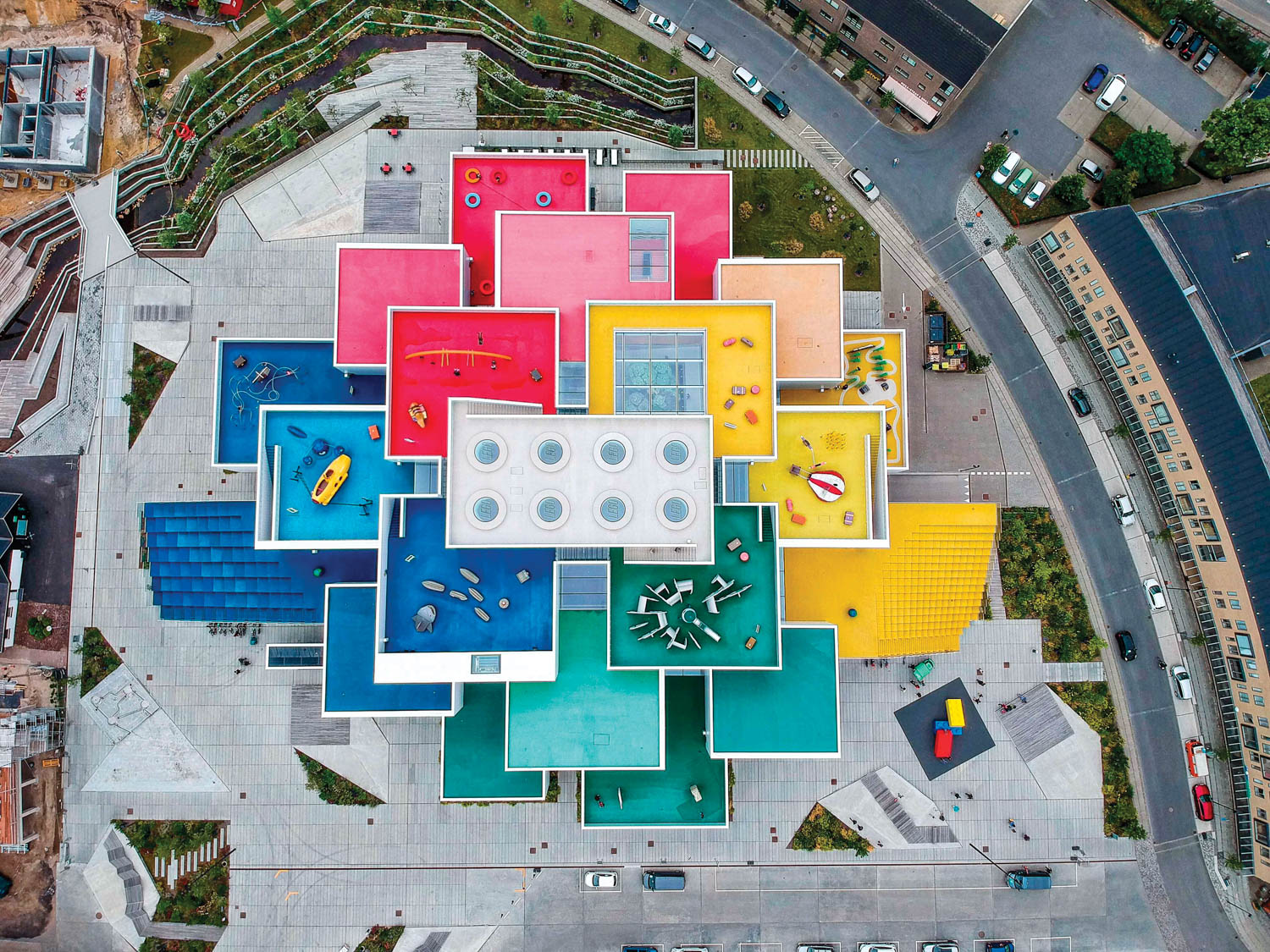 Aerial view of a colorful, block-shaped building complex with red, yellow, blue, and green roofs surrounded by roads and walkways.