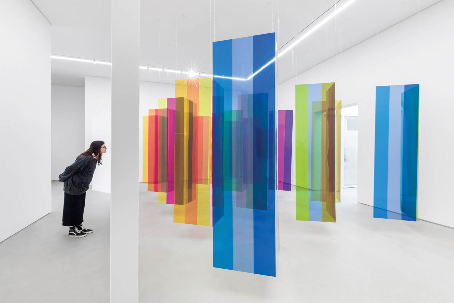 A person observes colorful, vertical transparent panels suspended from the ceiling in a bright, white art gallery.