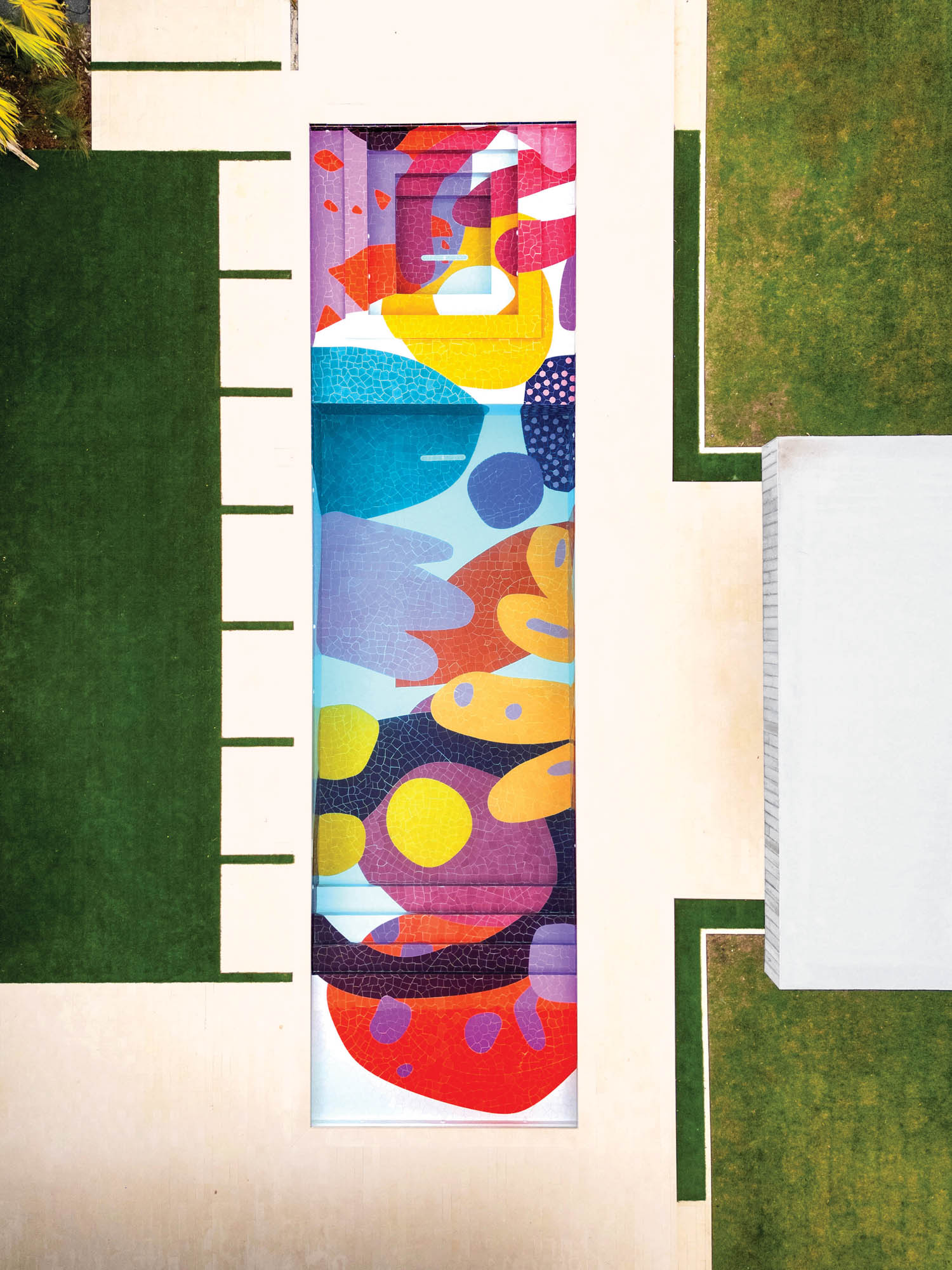 A rectangular swimming pool features a colorful abstract mural on its floor, surrounded by green grass and beige pavement, viewed from above.