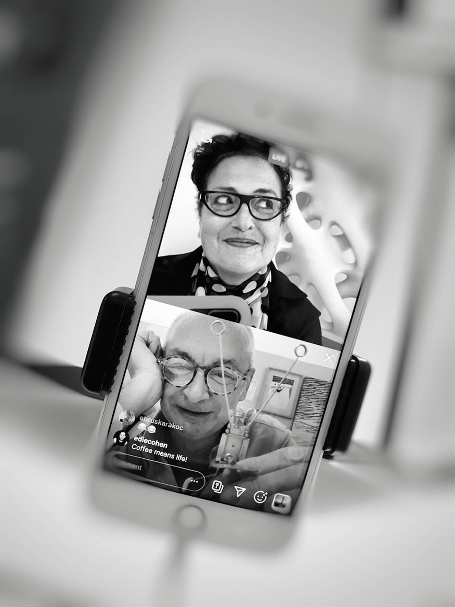 A smartphone displays a video call between a woman and a man, both smiling, in black and white. The phone is held upright in a stand.