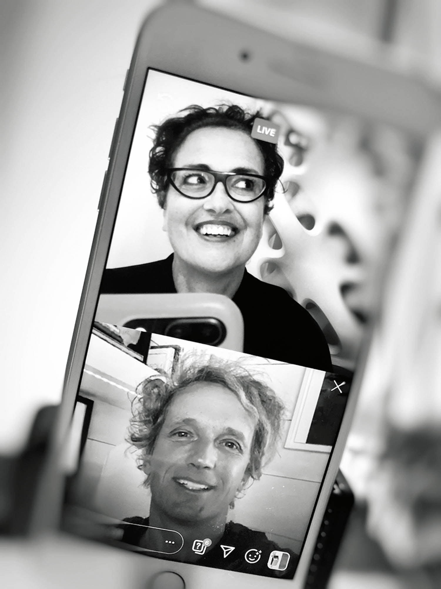 A smartphone screen shows a black and white video call with one person in the top half smiling and another person in the bottom half, both visible via front cameras.