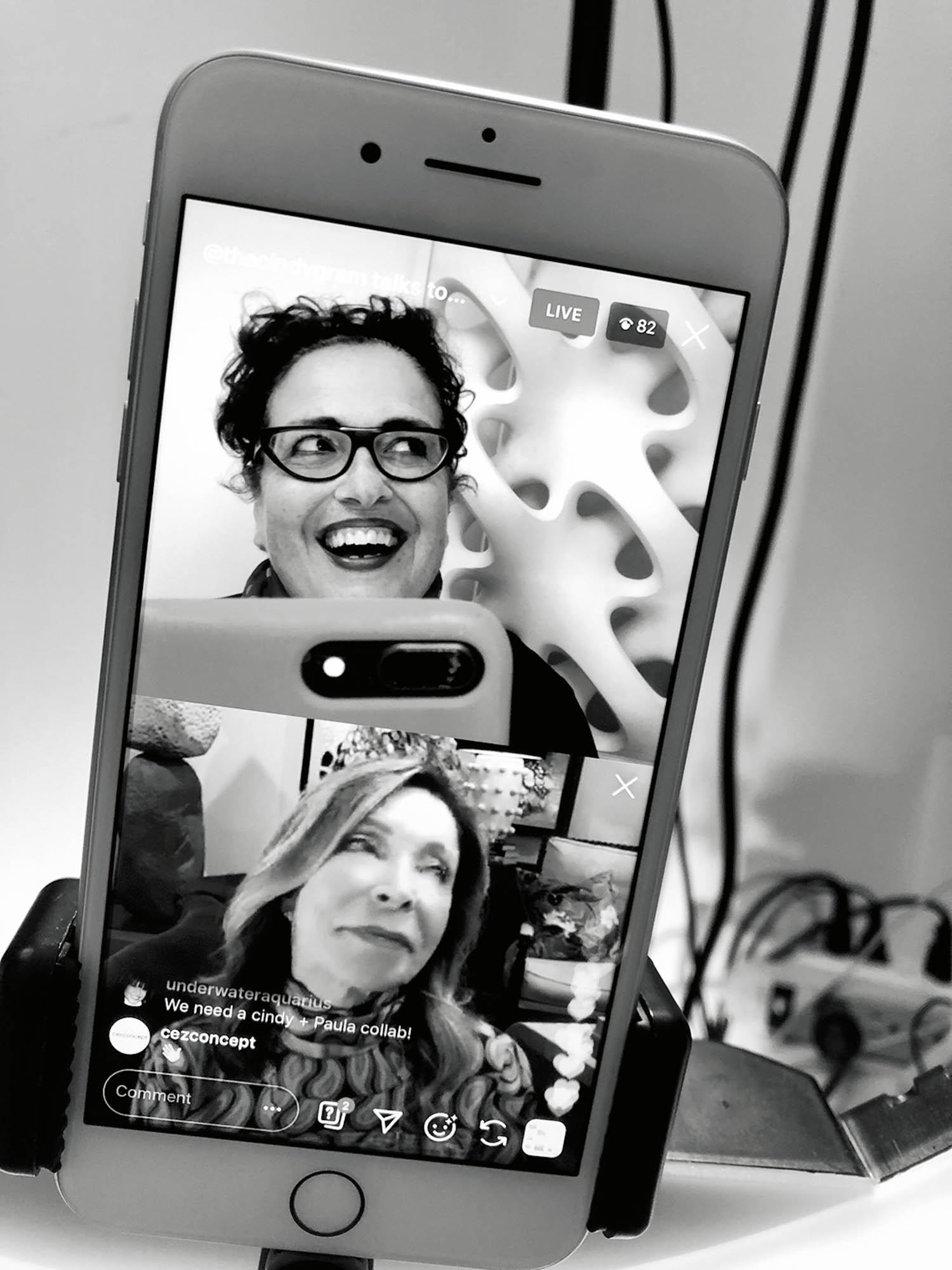 A phone displays a split-screen video call with two people smiling and laughing; the phone is mounted on a stand among cables and other electronic devices.