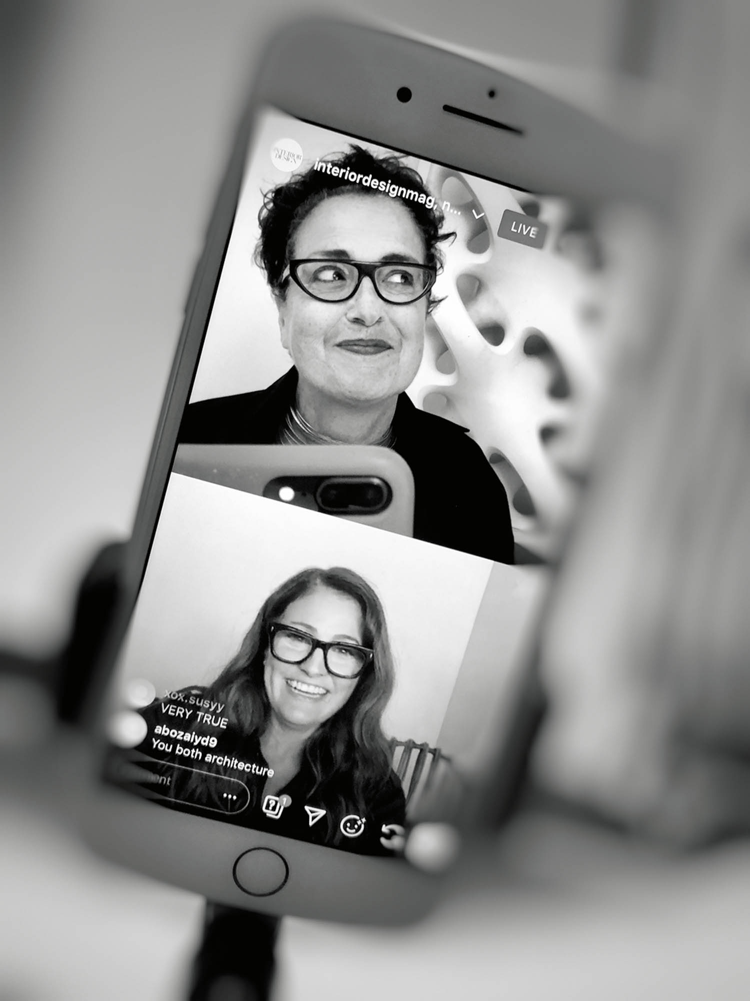 Black and white photo of a smartphone screen showing two women chatting during a live video call, both wearing glasses and smiling. The phone is mounted on a tripod.