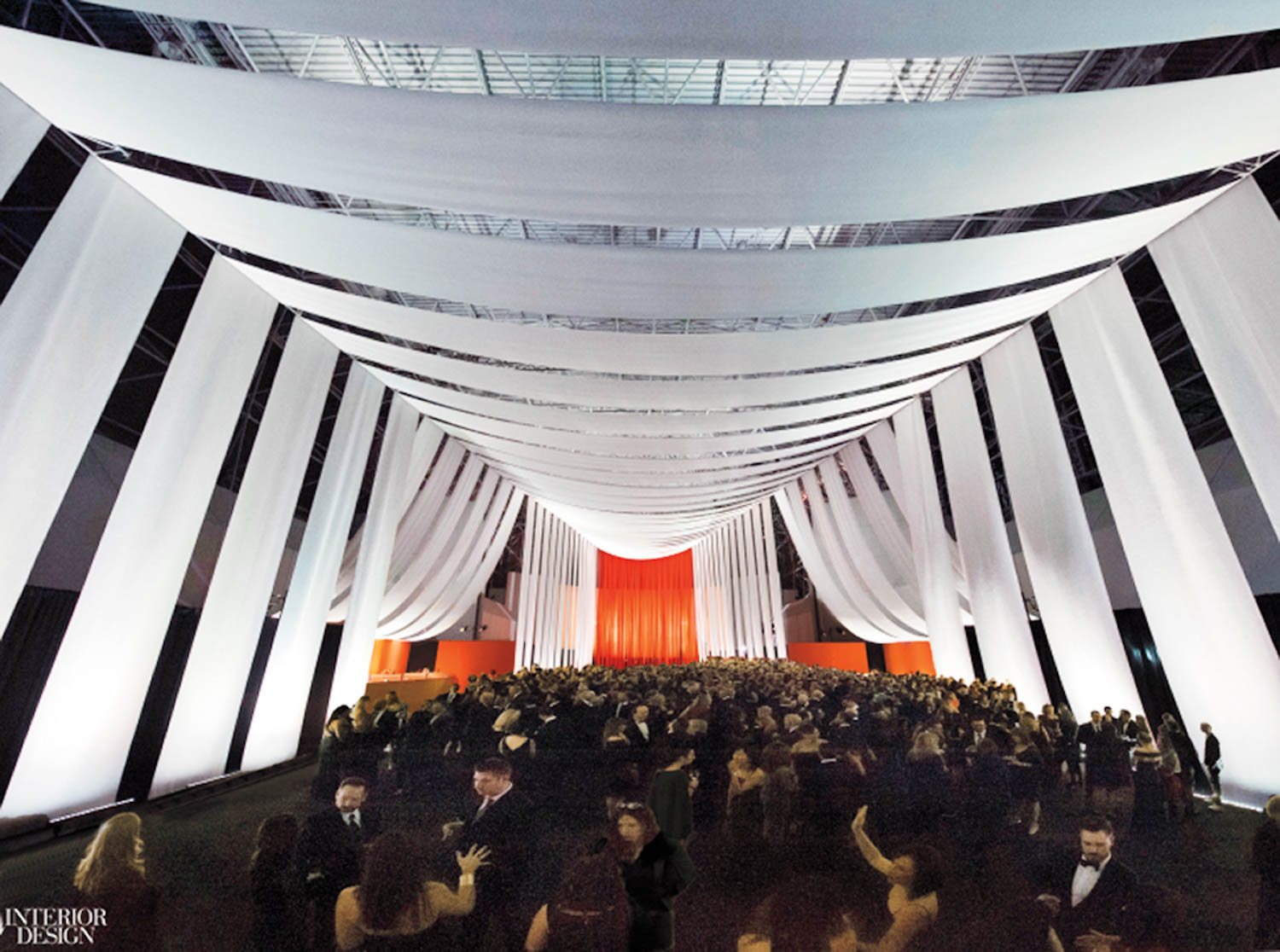 Large crowd gathers under dramatic white draped fabric panels in a spacious indoor venue with high ceilings and red backdrop.