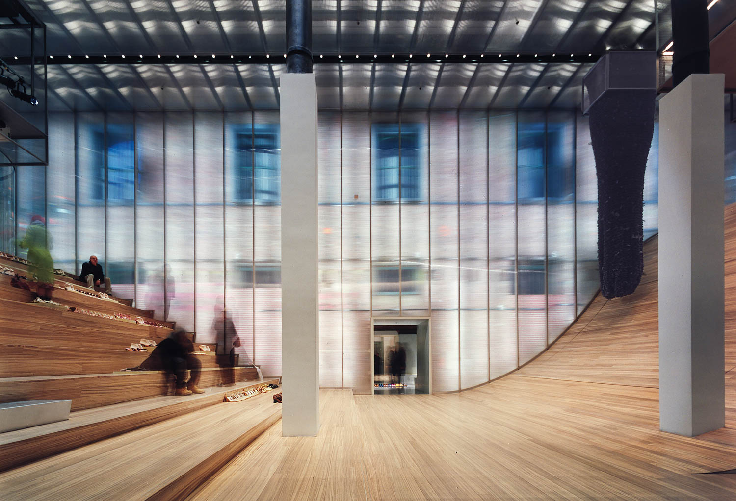 Modern interior with curved wooden seating, translucent wall revealing building facade behind, and a tall column in the center. A few people are seated on the steps.