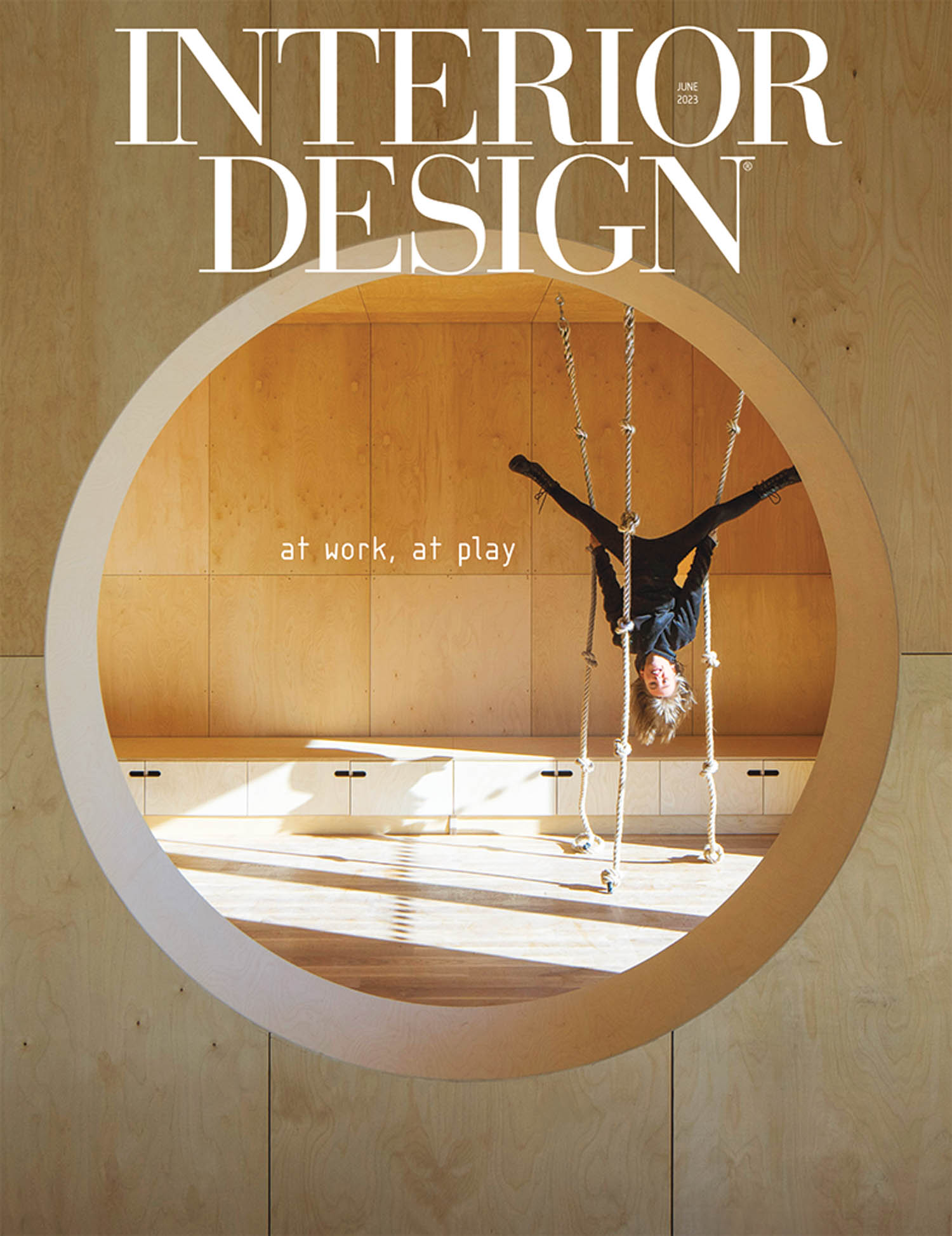 Person hangs upside down on rope swing inside a wooden room with circular opening; “Interior Design” magazine cover, June 2023, with text “at work, at play.”.