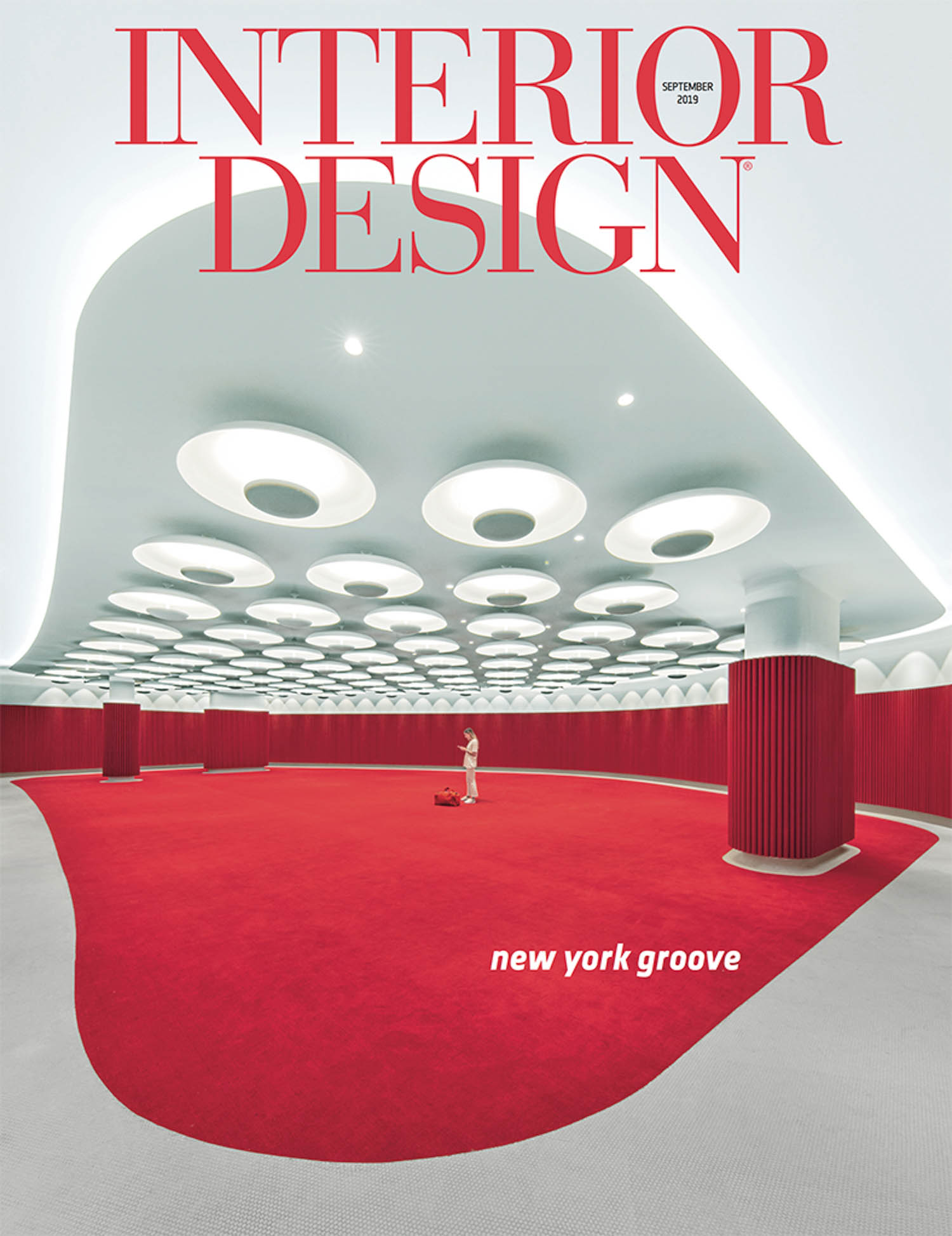 A person stands in a large red-carpeted room with circular ceiling lights and red columns. The magazine cover reads “INTERIOR DESIGN” and “new york groove.”.