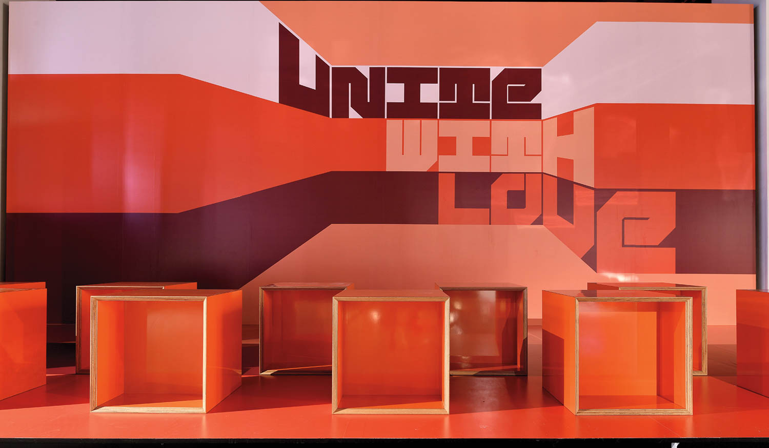 Orange and red geometric backdrop with the words “Unite With Love” and six open cube stools arranged in front.