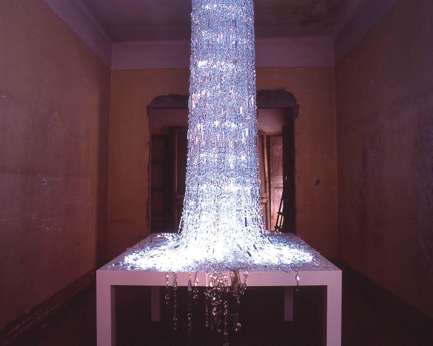 A cascade of sparkling, transparent strands flows from the ceiling to a white table, pooling on its surface, in a dim, empty room with yellowed walls and a doorway in the background.