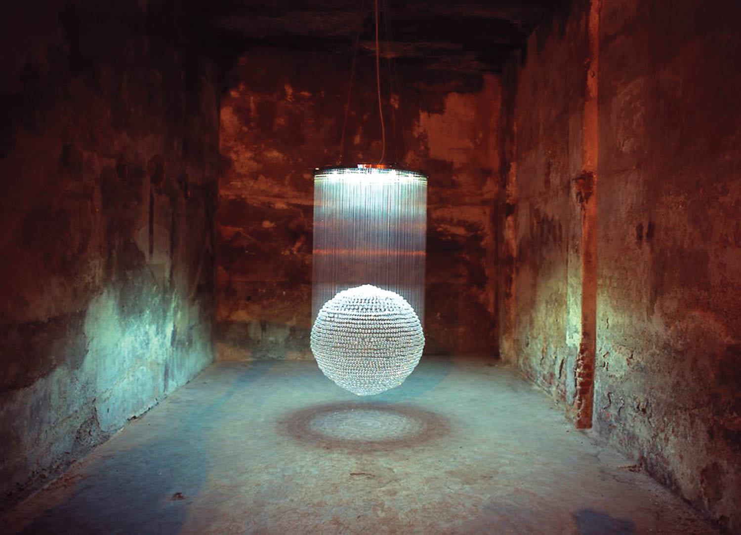 A suspended cylindrical light fixture with hanging strings and a glowing sphere in the center of a dim, empty, concrete room.
